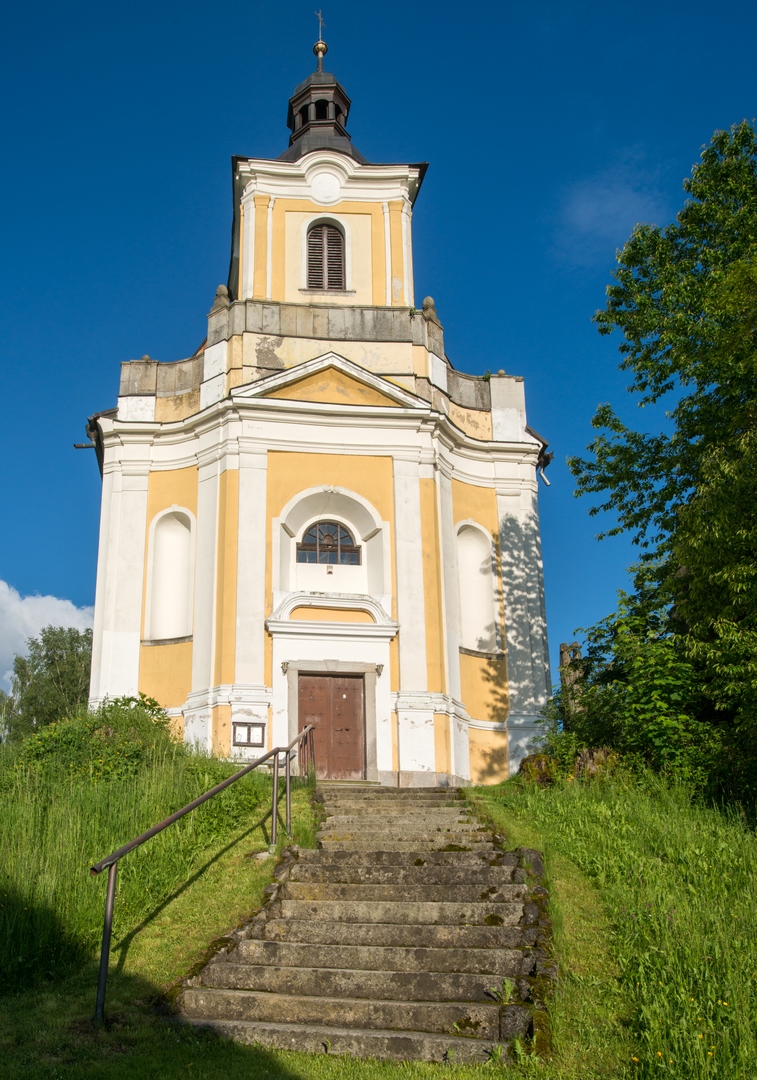 Barokní kostel s věží, schodištěm a dřevěnými dveřmi na pozadí modré oblohy a zeleně.